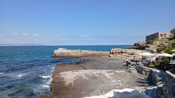 A sunny day, facing the sea with a rocky seafront and restaurant dining in the foreground.